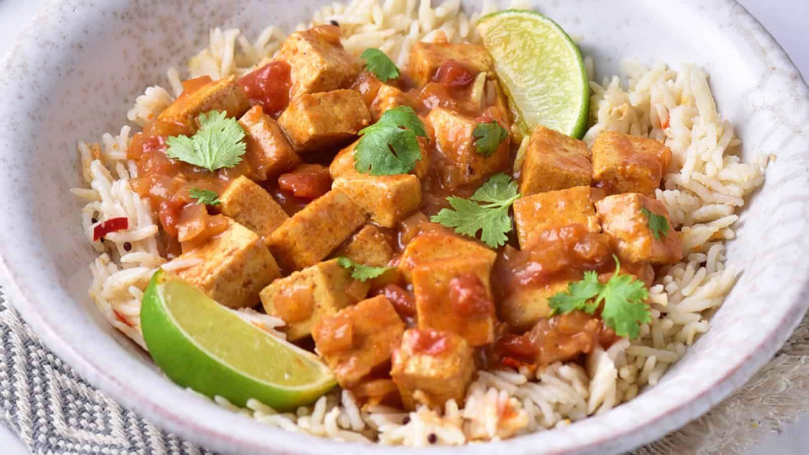 Tofu cubes coated in curry sauce with visible onions, served over fluffy rice and fresh herbs.