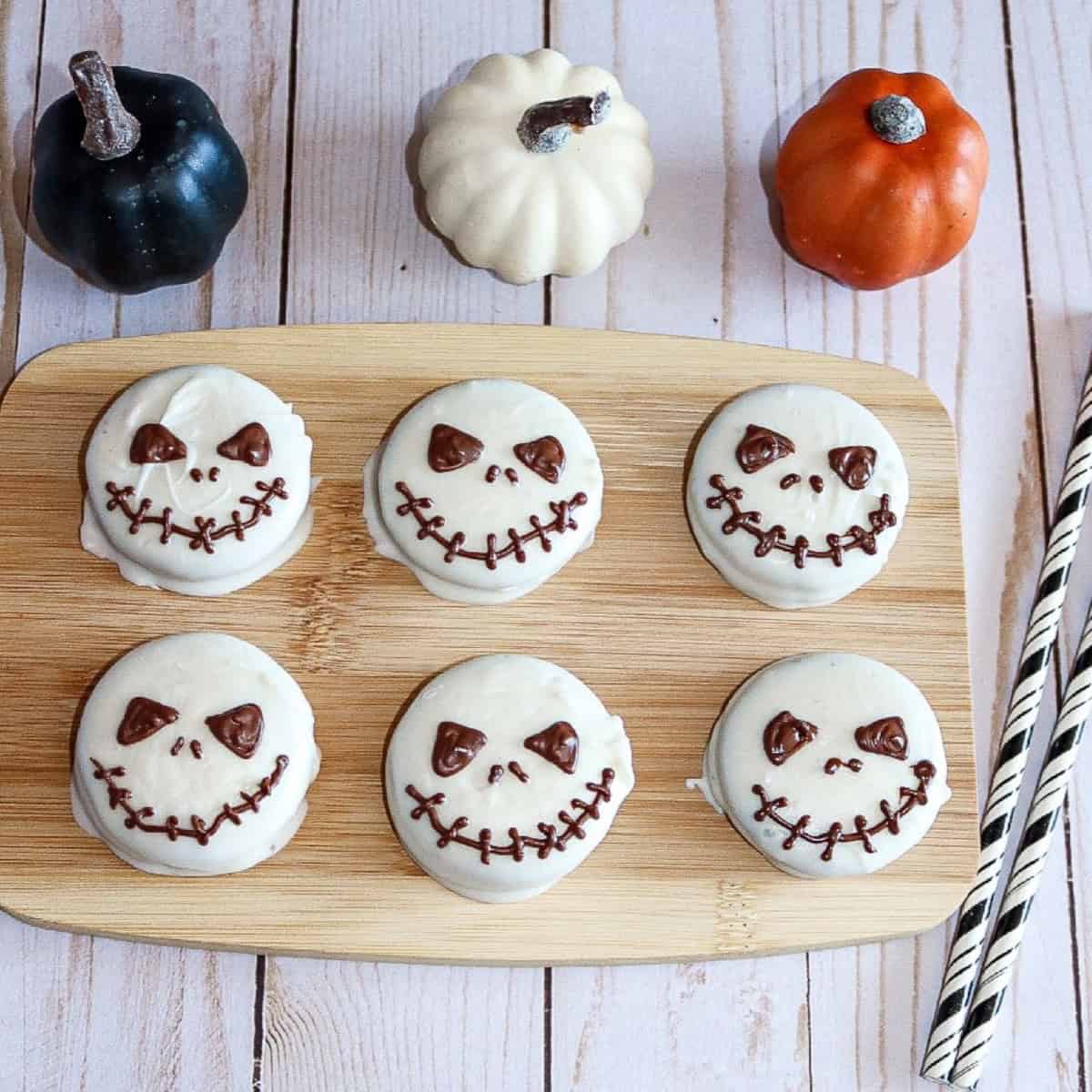 Halloween Jack Skellington Oreo cookies decorated with white chocolate and dark chocolate faces, displayed on a wooden board with mini pumpkins for festive decor.
