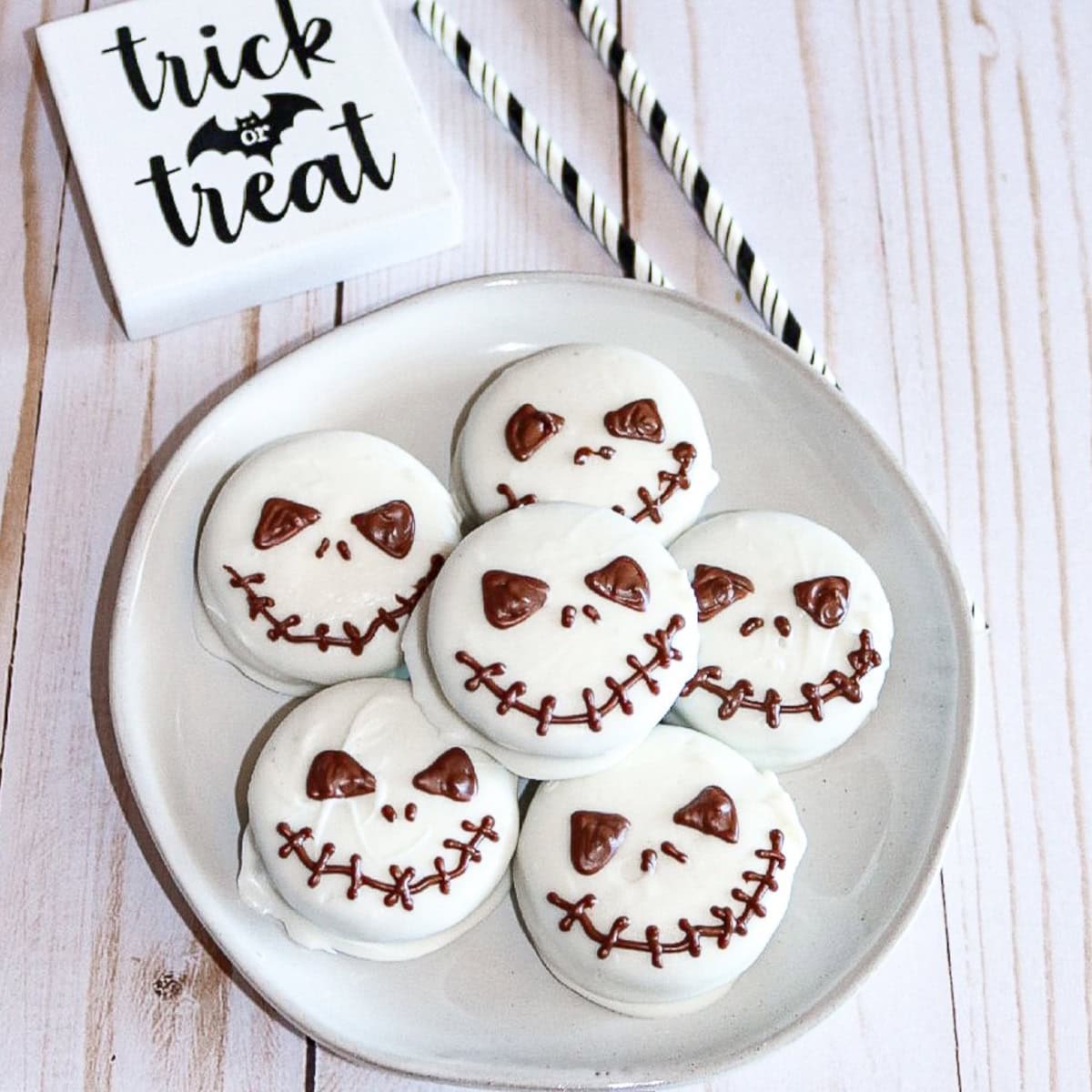 Plate of Jack Skellington Oreo cookies coated in white chocolate with spooky chocolate faces, styled with “Trick or Treat” decor and Halloween straws.