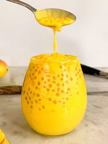 A spoon dripping creamy mango sago pudding into a glass filled with tapioca pearls and rich mango coconut mixture.