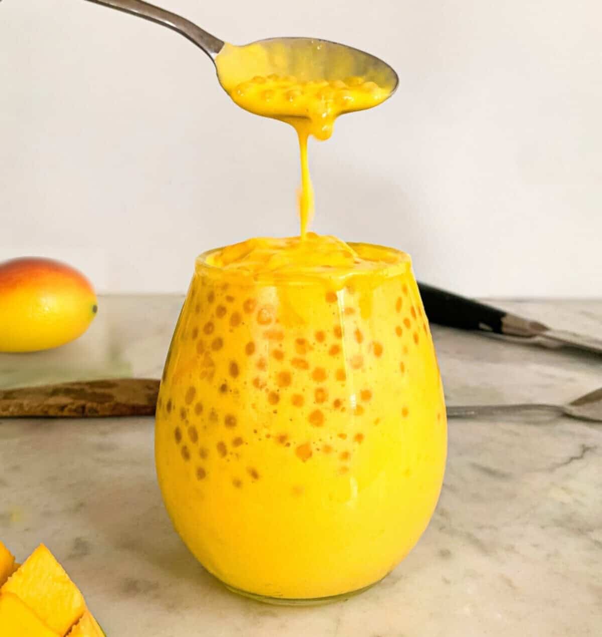 A spoon dripping creamy mango sago pudding into a glass filled with tapioca pearls and rich mango coconut mixture.