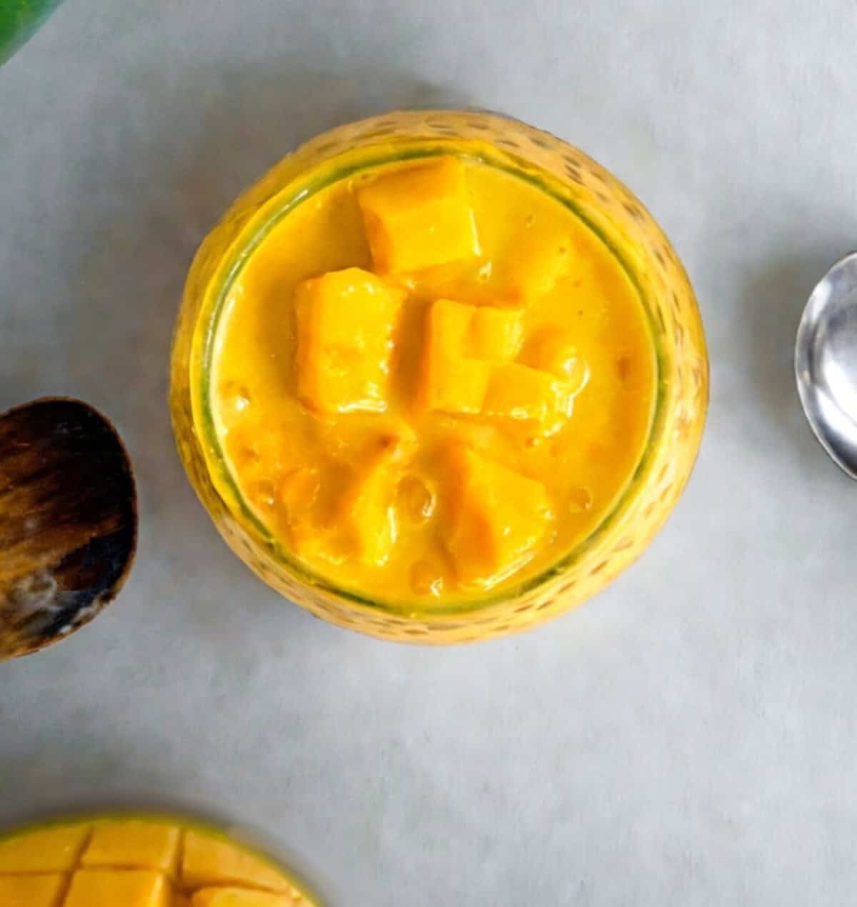 Top view of a glass filled with mango sago pudding and fresh mango chunks, beside a spoon and wooden spatula.