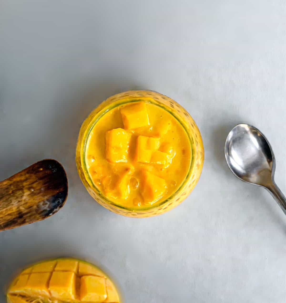Flat lay of creamy mango sago with diced mango, spoon, and wooden spatula on a gray background.