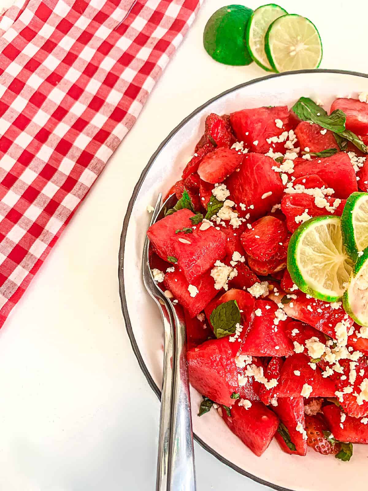 Close-up of spoon in a vibrant watermelon strawberry feta salad with fresh mint and lime, set against a red checkered cloth and lime slices.