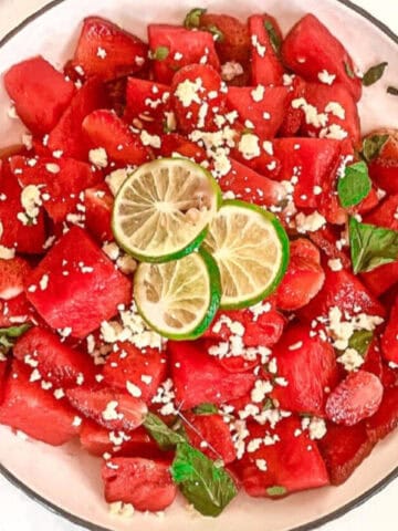 Top down view of watermelon strawberry salad with feta cheese, mint, and lime in a white bowl beside lime slices.