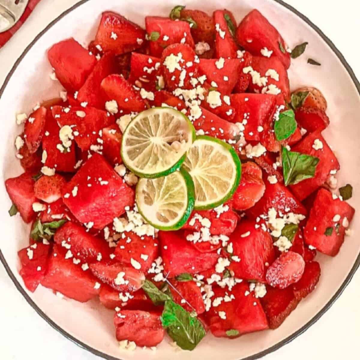 Top down view of watermelon strawberry salad with feta cheese, mint, and lime in a white bowl beside lime slices.