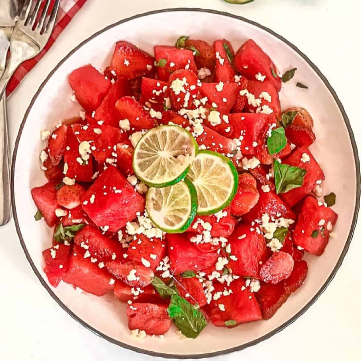 Top-down view of watermelon strawberry salad with feta, mint, and lime in a white bowl beside lime slices.