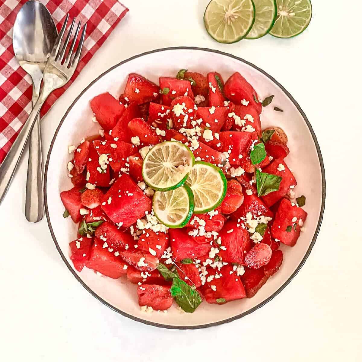 Top-down view of watermelon strawberry salad with feta, mint, and lime in a white bowl beside lime slices and silverware on a white table.