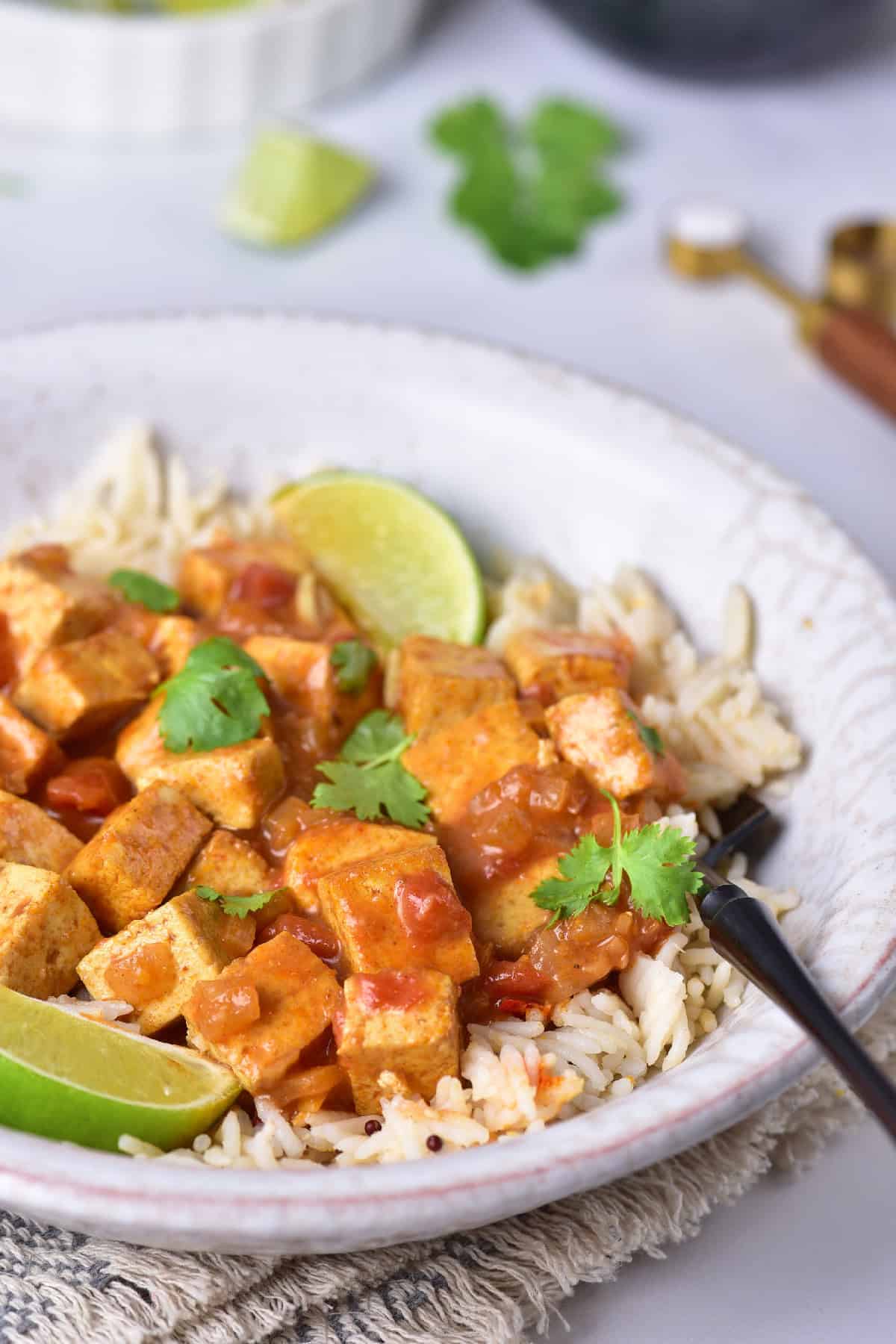 Close-up of creamy yellow tofu curry with rice, garnished with fresh cilantro and lime in a rustic bowl.