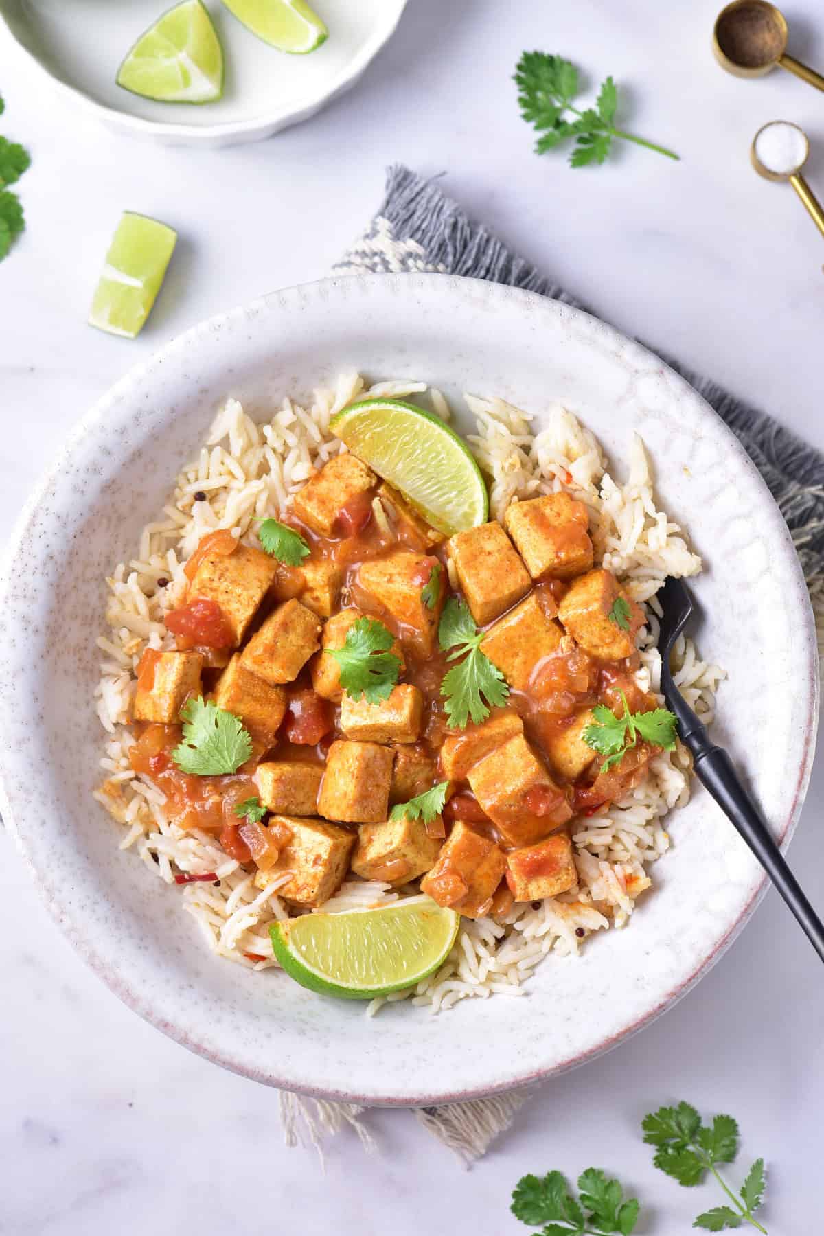 Overhead view of tofu curry served with rice and lime wedges, surrounded by scattered herbs on a white surface.