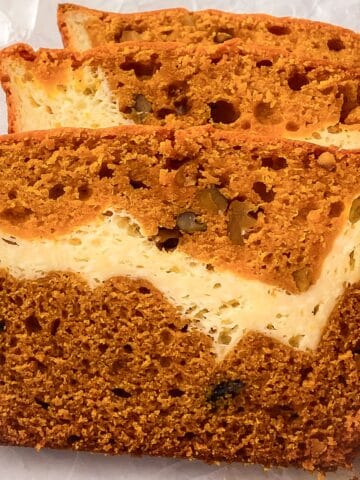 Close-up of moist cream cheese pumpkin bread slices showing a rich orange pumpkin layer swirled with creamy cheesecake filling and bits of nuts on a parchment-lined surface.