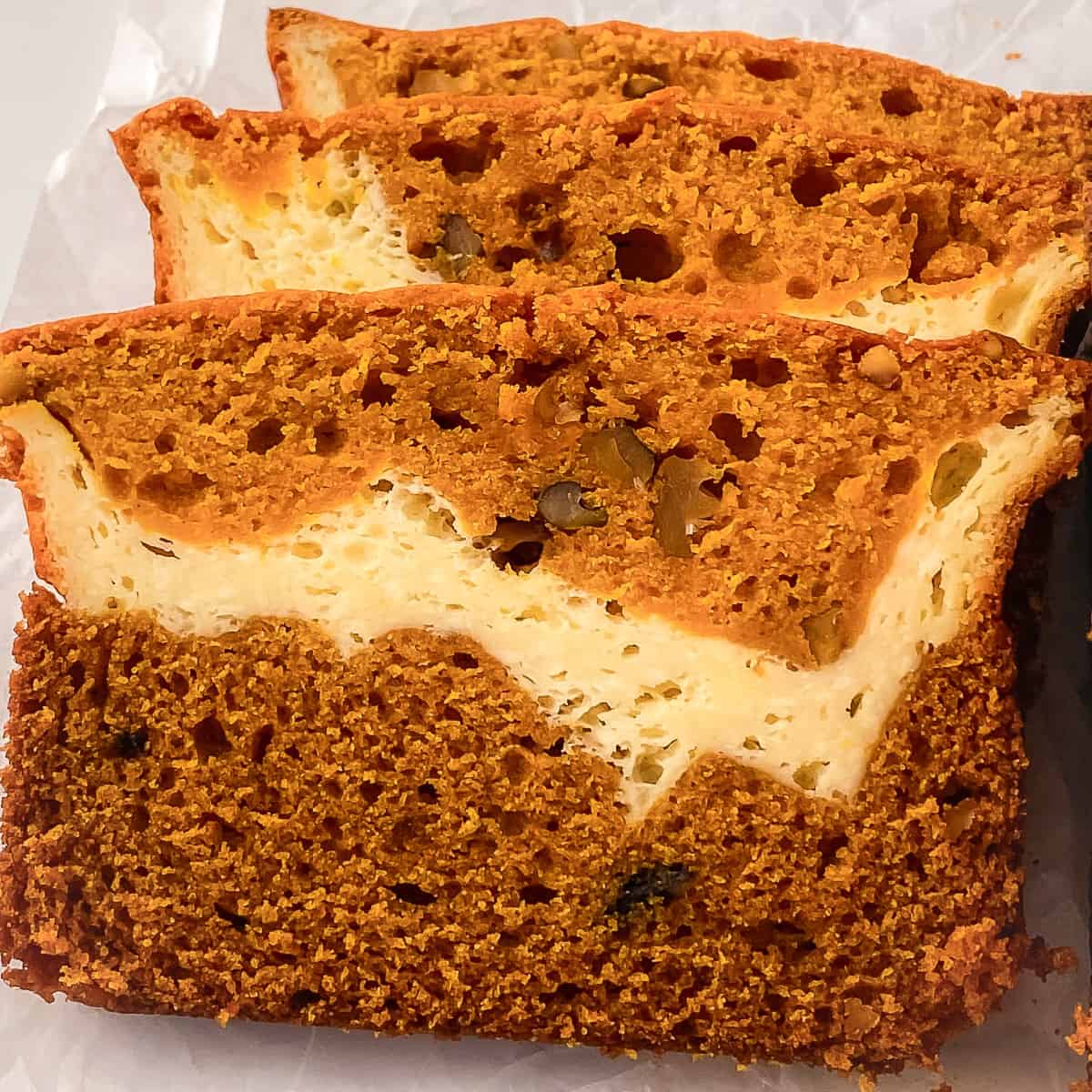 Close-up of moist cream cheese pumpkin bread slices showing a rich orange pumpkin layer swirled with creamy cheesecake filling and bits of nuts on a parchment-lined surface.