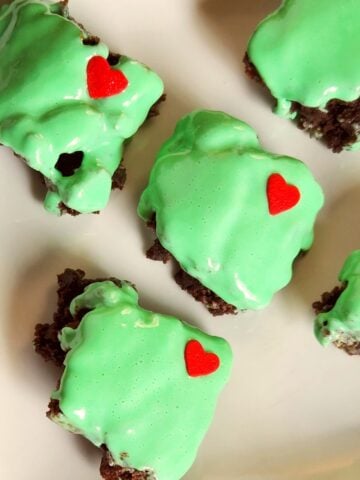 Small square chocolate brownies topped with bright green frosting and decorated with little red candy hearts, arranged on a white plate for a festive Grinch-inspired treat.
