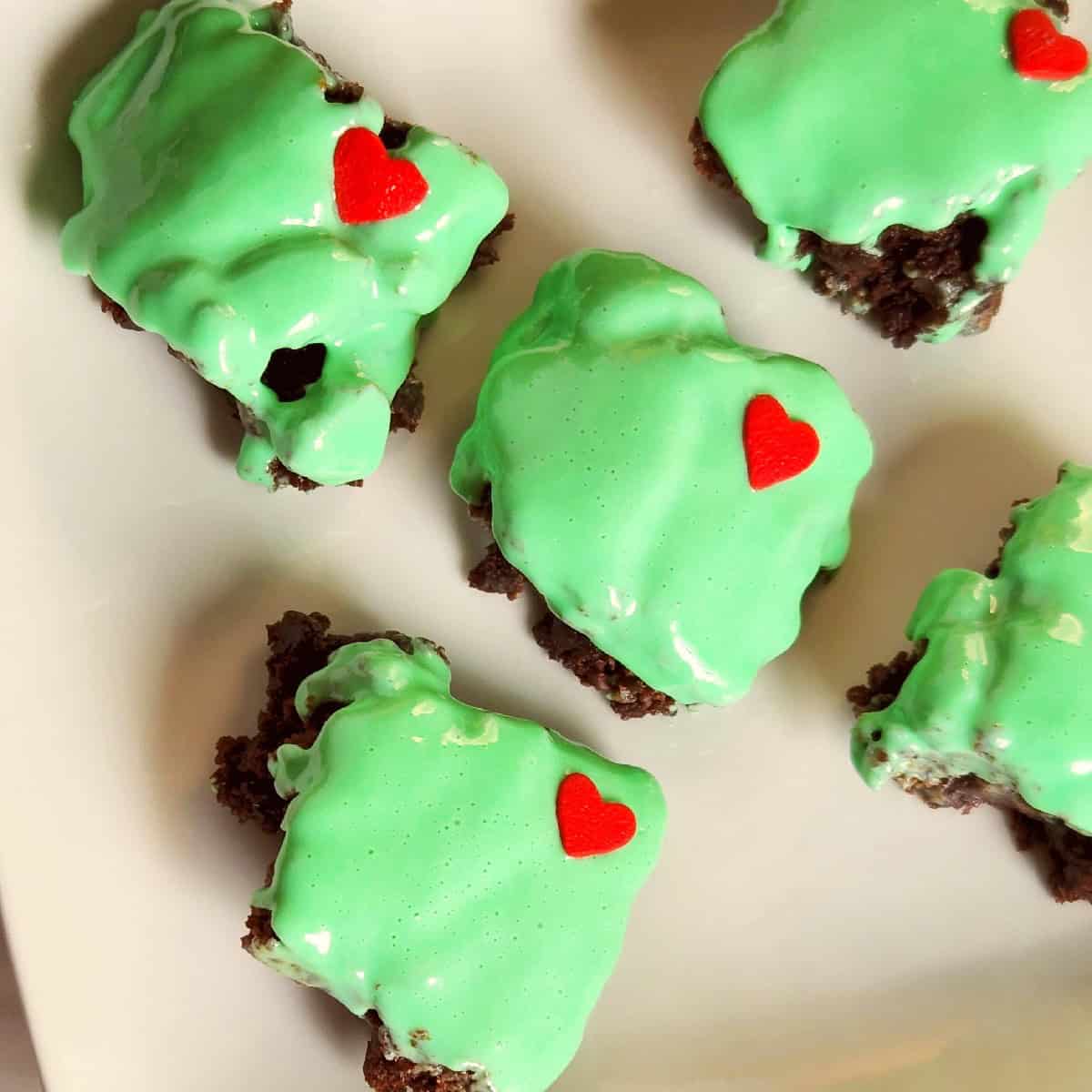 Small square chocolate brownies topped with bright green frosting and decorated with little red candy hearts, arranged on a white plate for a festive Grinch-inspired treat.