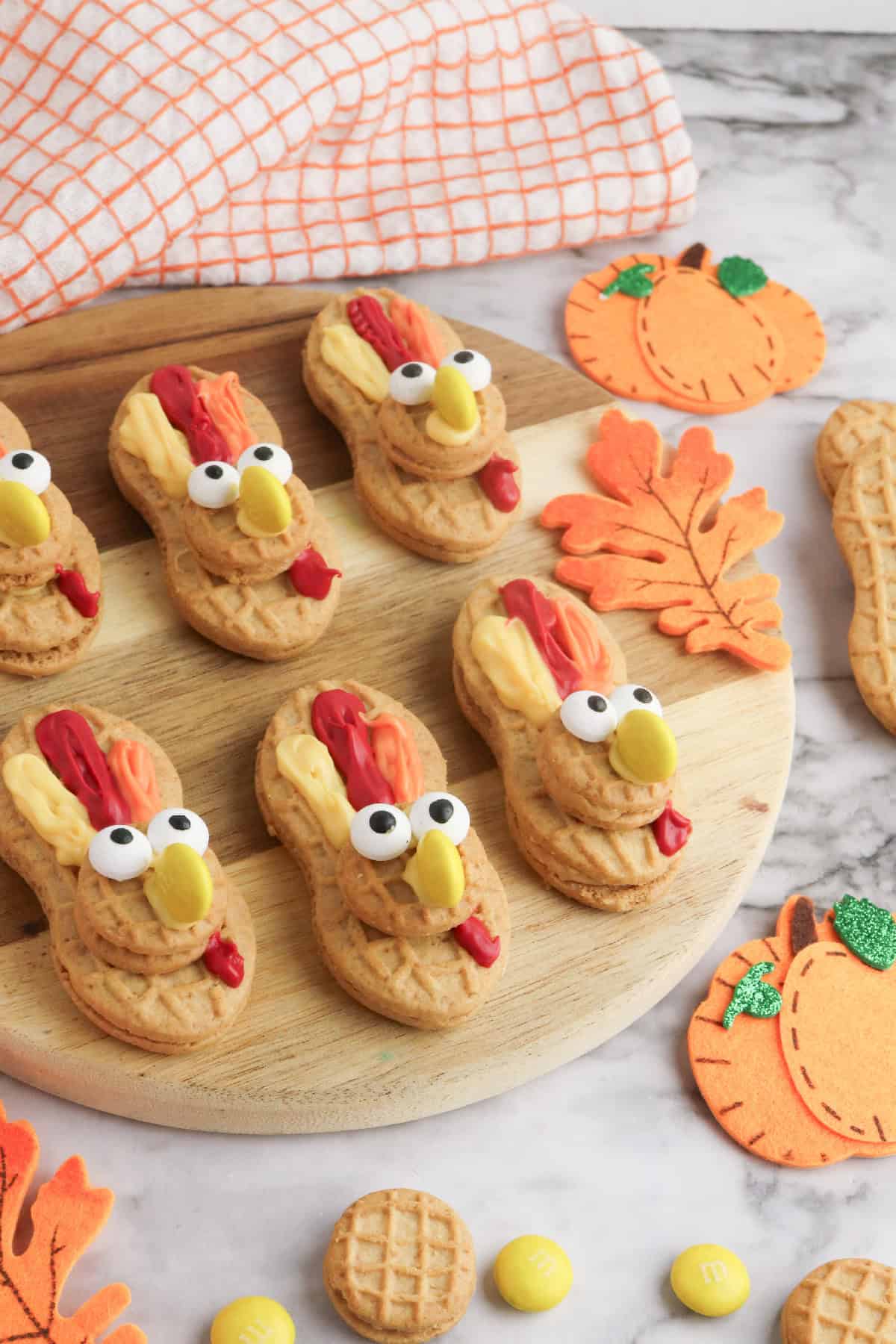 Homemade Nutter Butter turkey cookies decorated with candy eyes, yellow M&M beaks, and colorful icing feathers on a round wooden board with fall decorations.