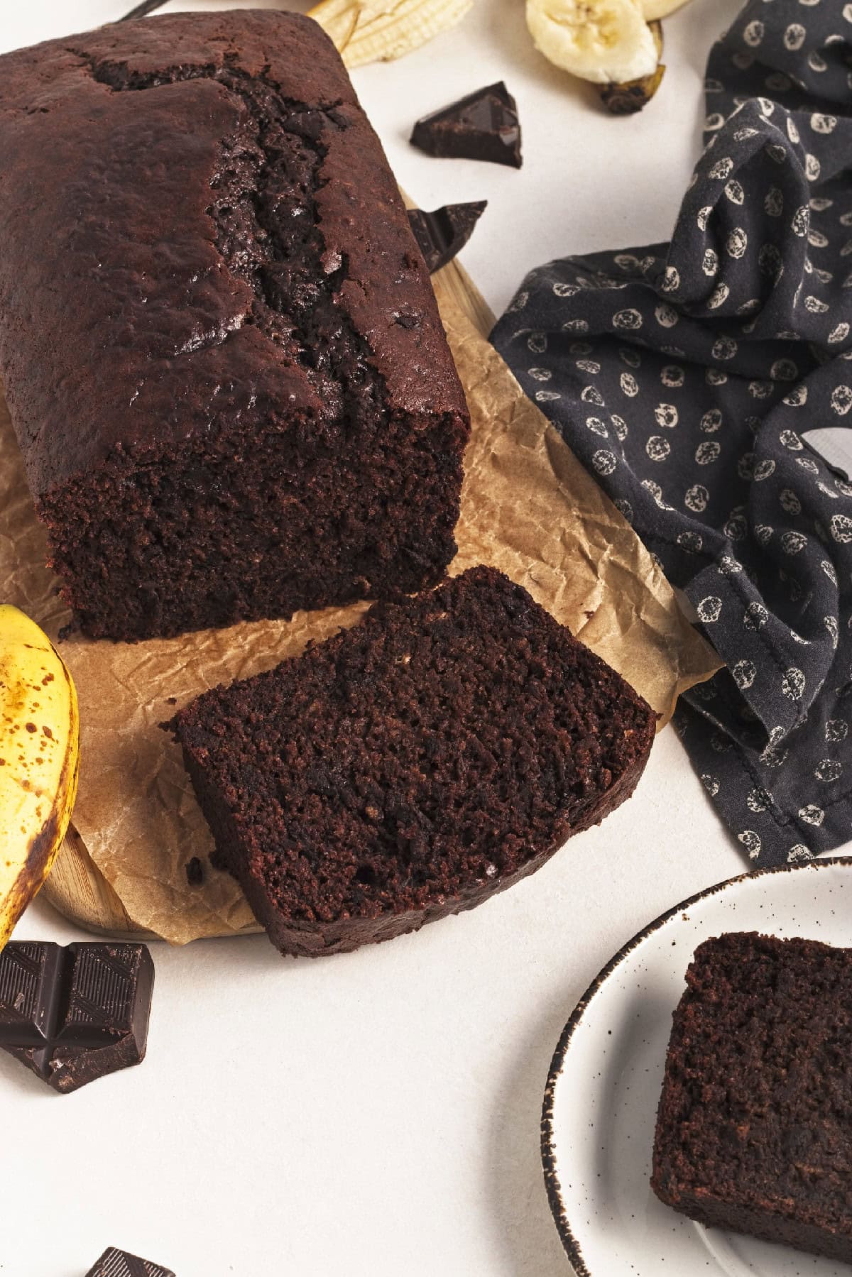 Angled shot of vegan chocolate banana bread loaf with one slice cut, banana and chocolate pieces on the side.