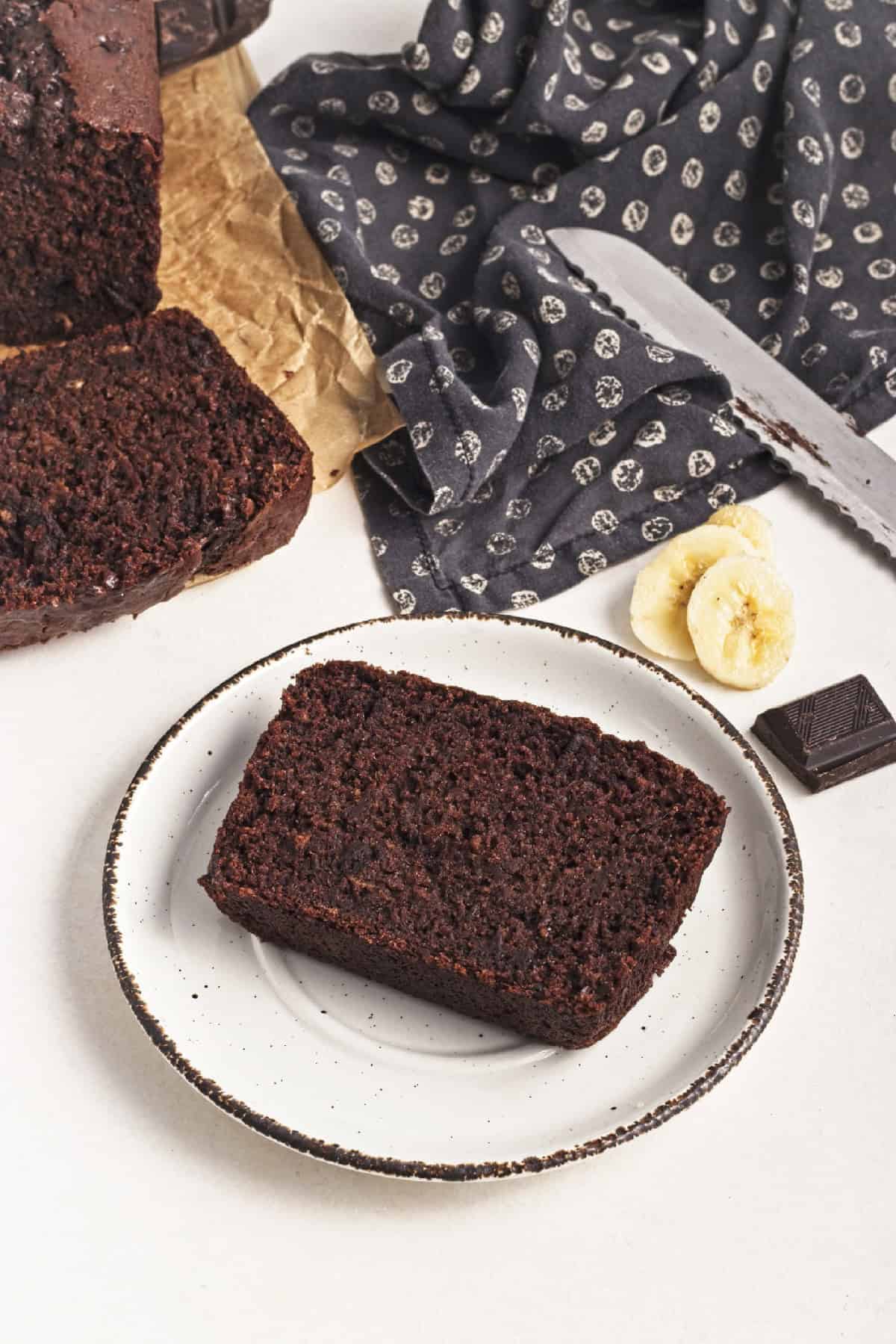 Close-up of a slice of vegan chocolate banana bread on a white plate with banana and chocolate garnish.