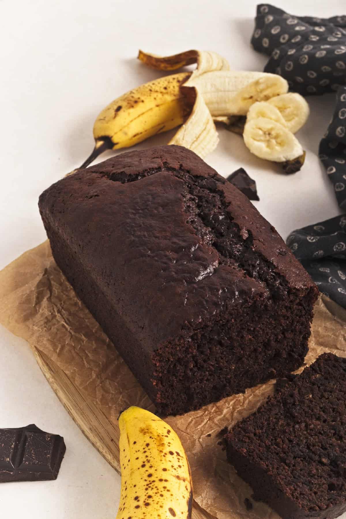 Vegan chocolate banana bread loaf cooling on parchment with banana peel and chocolate pieces in the background.