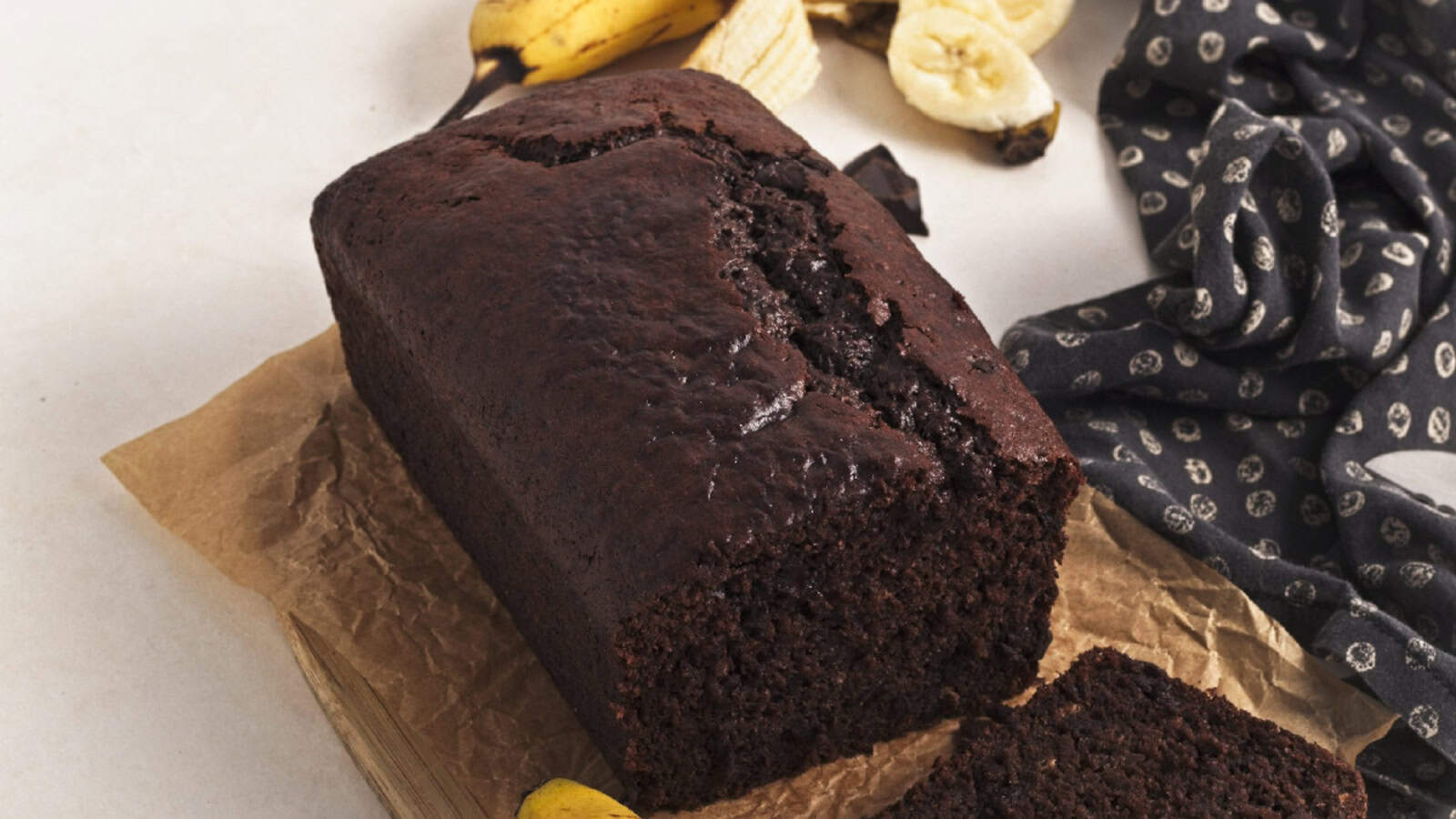 Vegan chocolate banana bread loaf cooling on parchment paper with banana peel and chocolate pieces in the background.