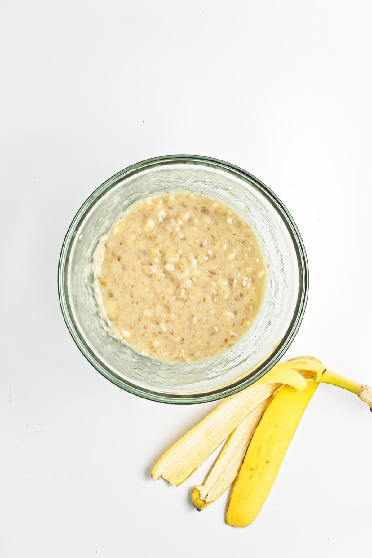 Mashed ripe bananas in a glass mixing bowl with banana peel beside it – base for vegan chocolate banana bread.