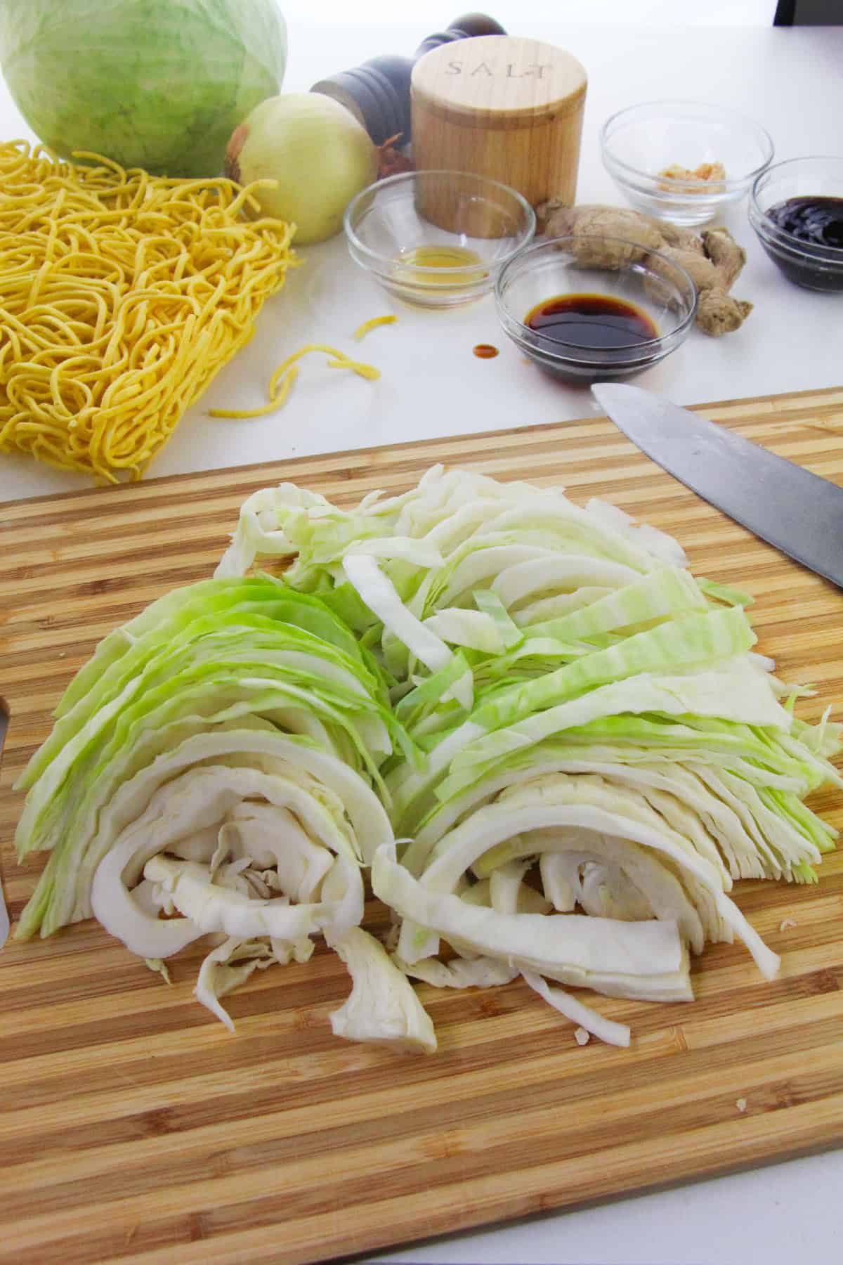Fresh cabbage sliced on a wooden cutting board with yellow noodles, onion, ginger, and small bowls of soy sauce, oil, and seasonings in the background, ready for making chow mein.