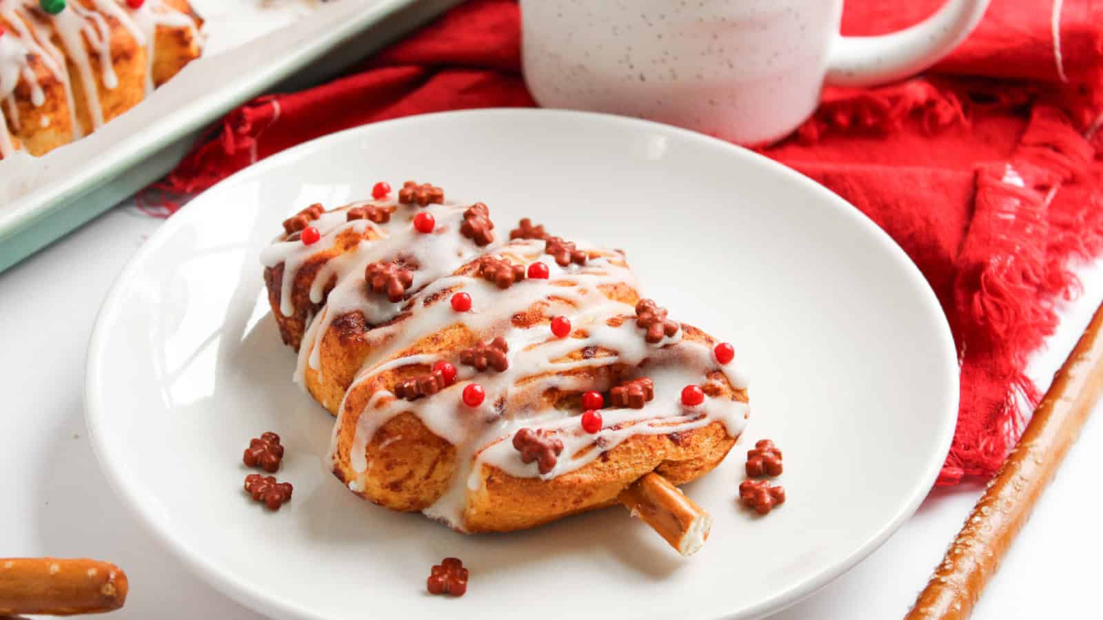 Christmas tree cinnamon roll decorated with white icing and festive sprinkles, served on a white plate with a red holiday cloth in the background.