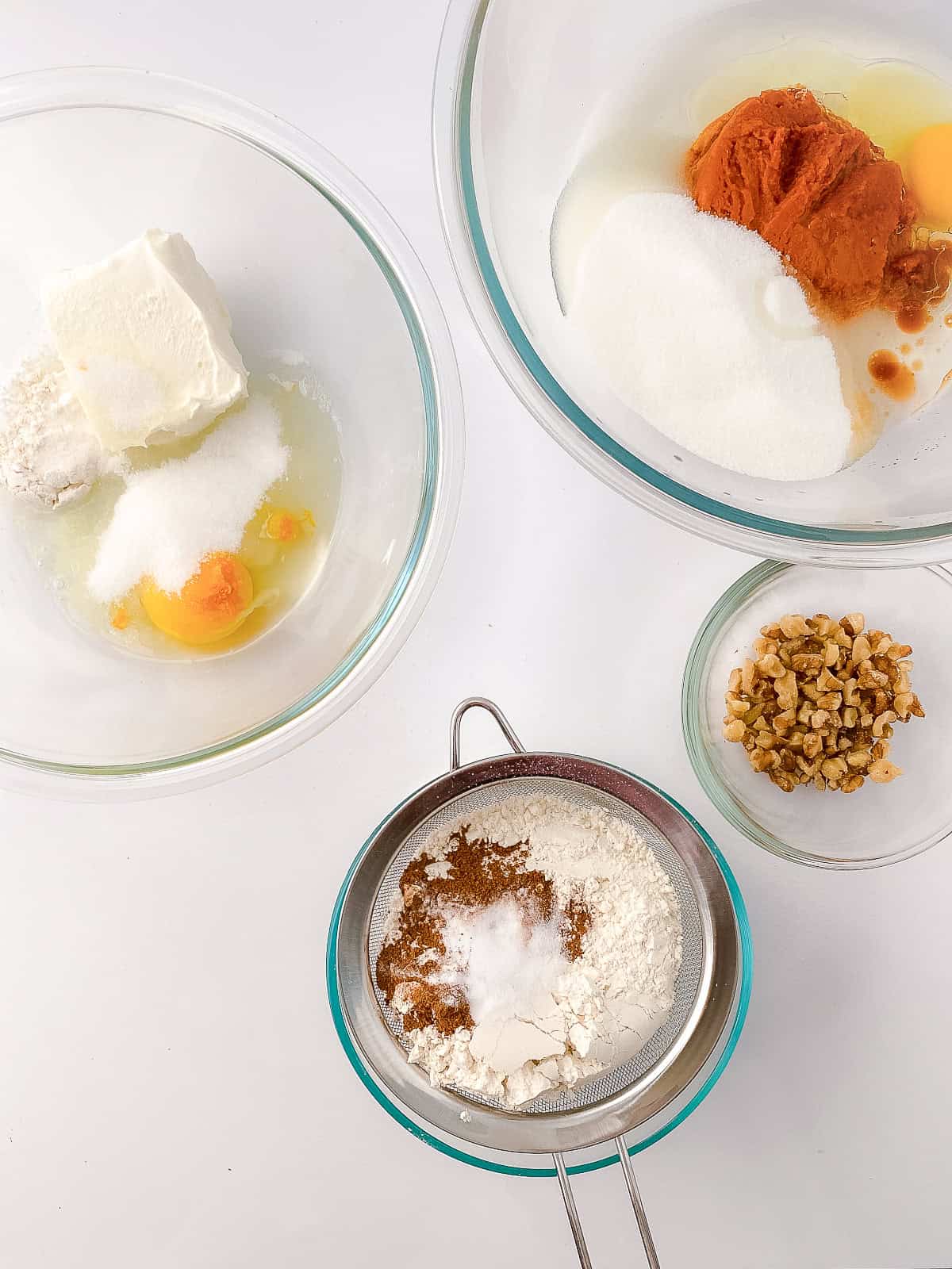 Ingredients for cream cheese pumpkin bread laid out in glass bowls on a white surface - softened cream cheese, egg, sugar, flour, pumpkin purée, vanilla, baking spices in a sieve, and chopped walnuts.