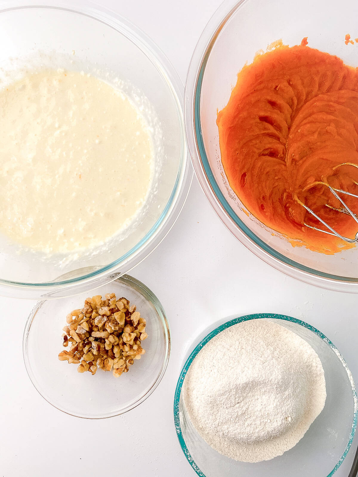 Mixed cream cheese filling and pumpkin batter in glass bowls with chopped walnuts and sifted dry ingredients arranged on a white surface for making cream cheese pumpkin bread.