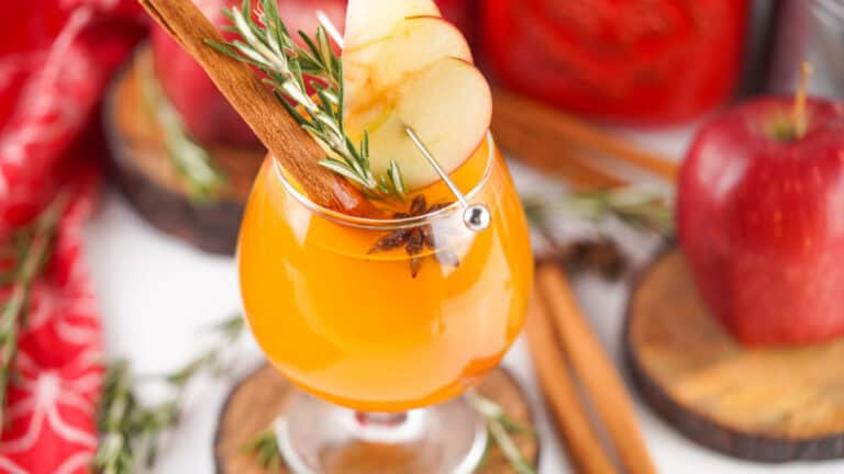 A glass of Fireball apple cider garnished with apple slices, a cinnamon stick, and rosemary, with apples and cinnamon sticks in the background.