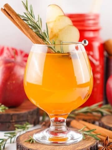 A festive glass of Fireball apple cider garnished with apple slices, a cinnamon stick, and rosemary, set against a cozy fall background with apples and spices.