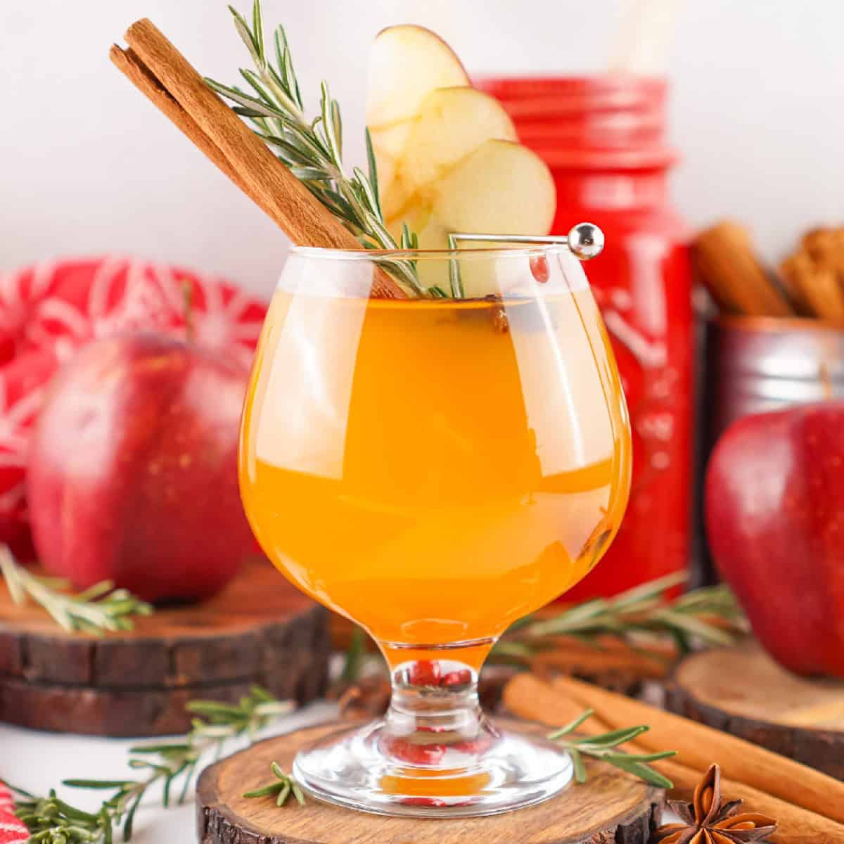A festive glass of Fireball apple cider garnished with apple slices, a cinnamon stick, and rosemary, set against a cozy fall background with apples and spices.