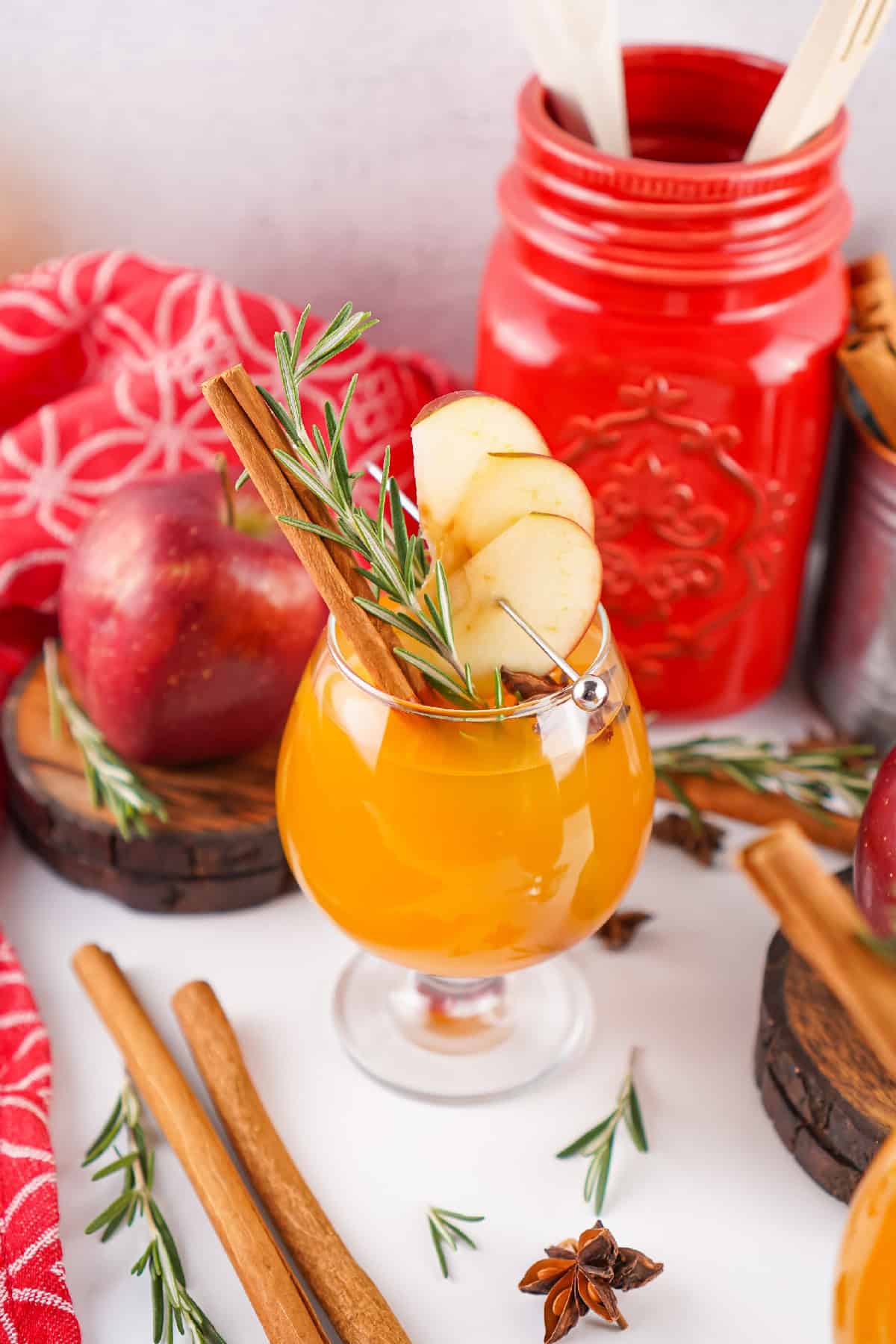 Close-up angled shot of Fireball apple cider with apple slices, rosemary, and star anise garnish, with red and festive background elements.