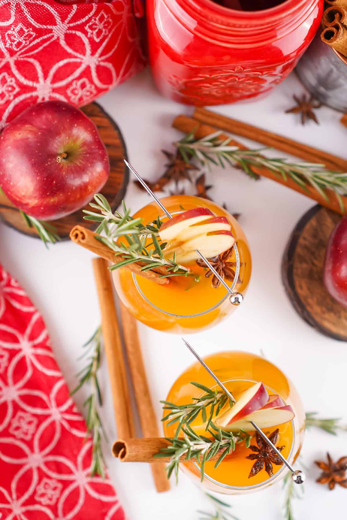 Two glasses of Fireball apple cider with apple slices, rosemary, and cinnamon sticks, styled with apples and a red holiday jar in the background.