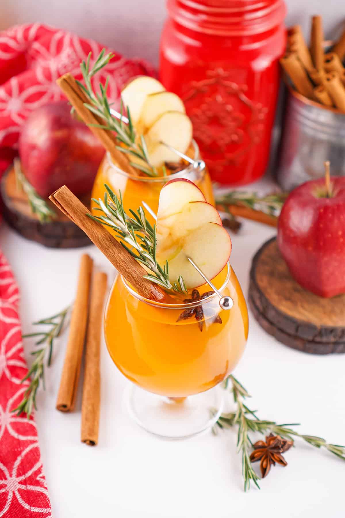 Festive scene with two Fireball apple cider cocktails, garnished with apple slices and rosemary, surrounded by apples, cinnamon sticks, and rustic props.