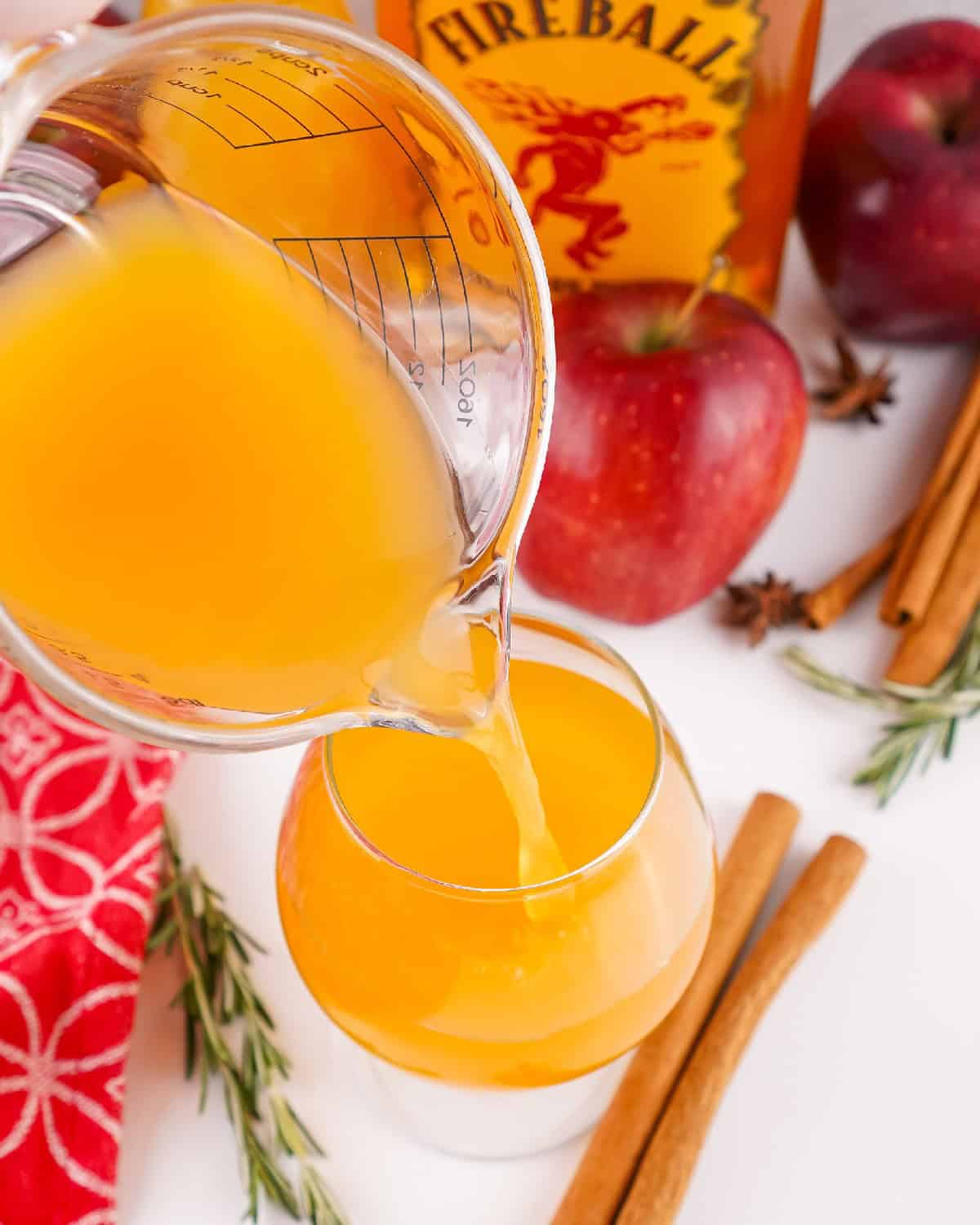 Apple cider being poured from a glass measuring cup into a round cocktail glass with apples and cinnamon in the background.