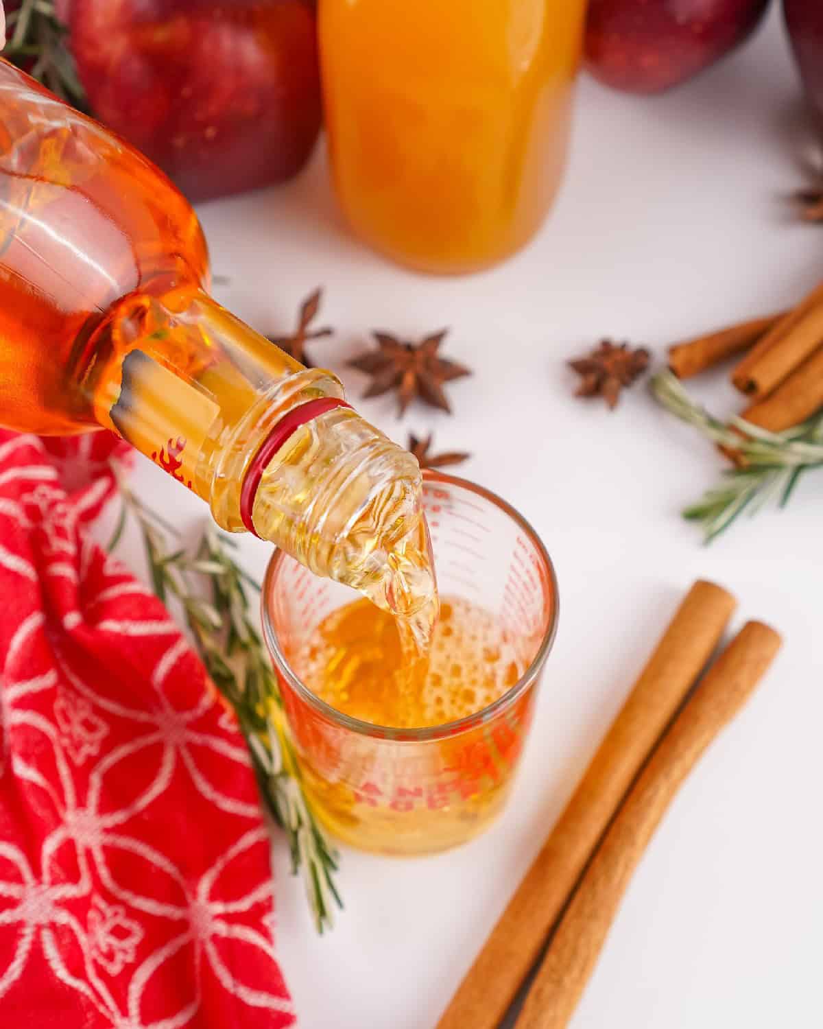 Fireball cinnamon whisky being poured from the bottle into a measuring cup with apples, cinnamon sticks, and cider in the background.