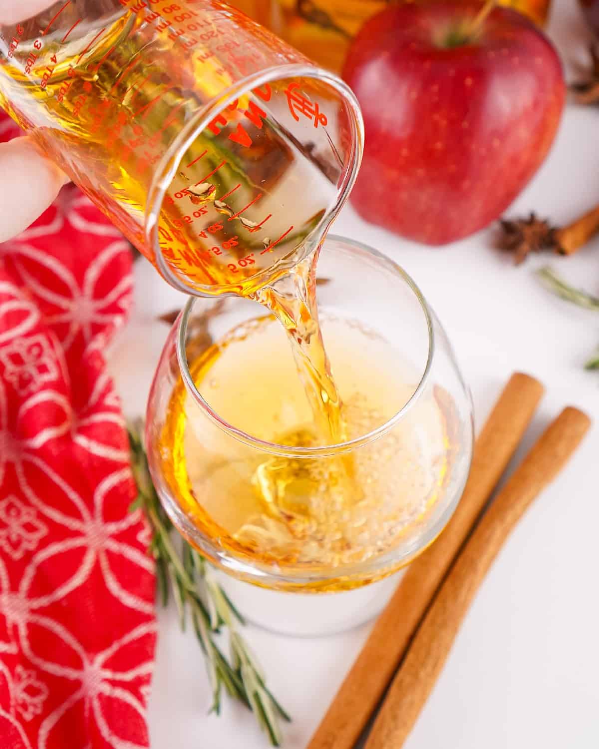 Golden Fireball whisky being poured from a measuring cup into a cocktail glass with apples, cider, and spices on the table.