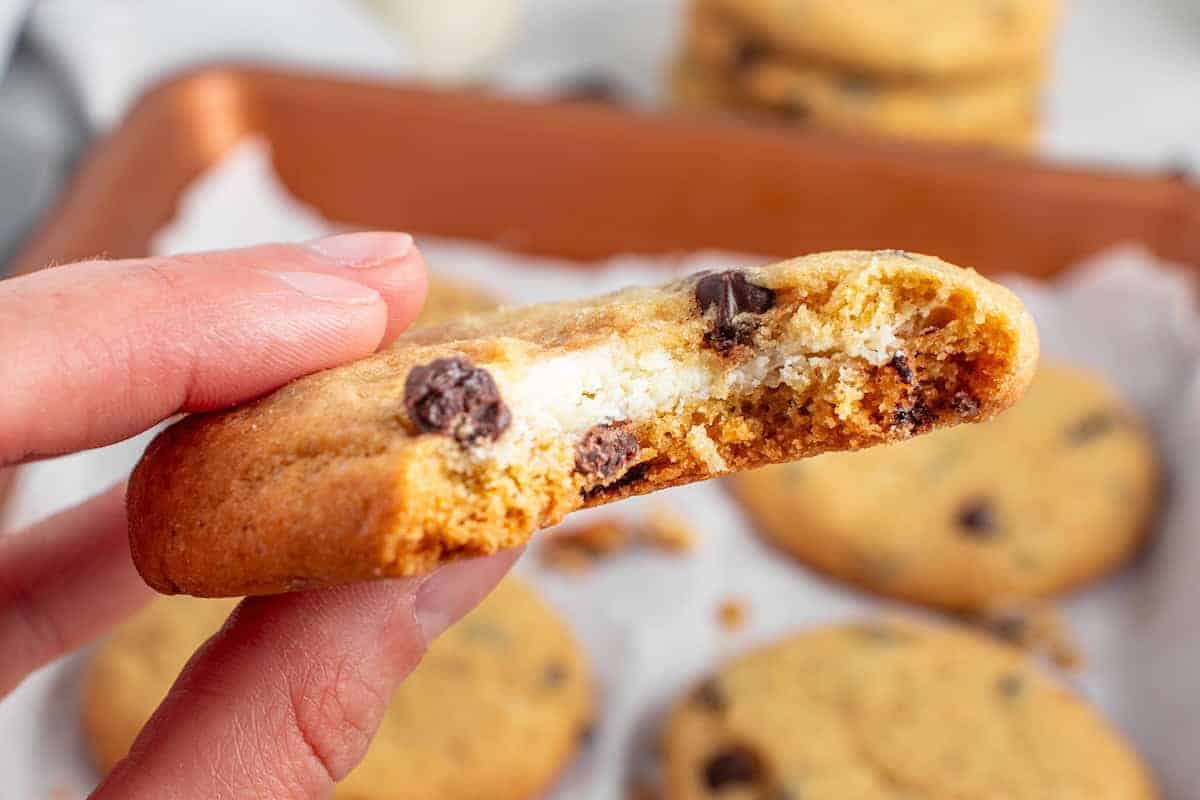A hand holding a half-eaten chocolate chip cookie with a cheesecake filling. Other cookies are visible on a baking tray in the background.