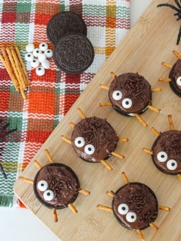 Halloween Oreo spider cookies with candy eyes and pretzel legs arranged on a wooden board with toy spiders and fall napkin.
