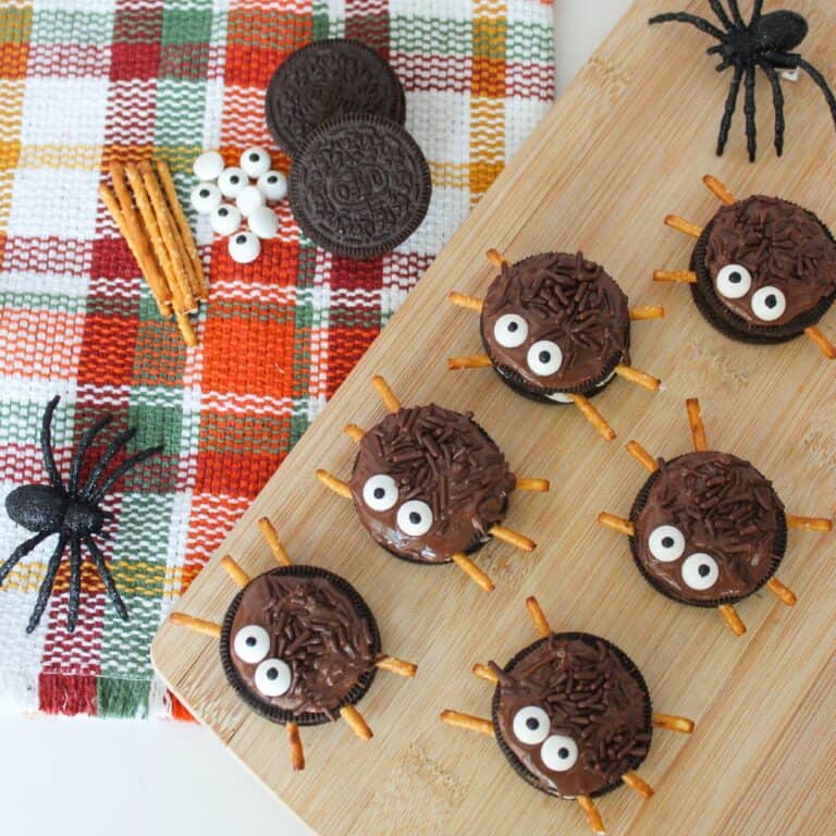 Halloween Oreo spider cookies with candy eyes and pretzel legs arranged on a wooden board with toy spiders and fall napkin.
