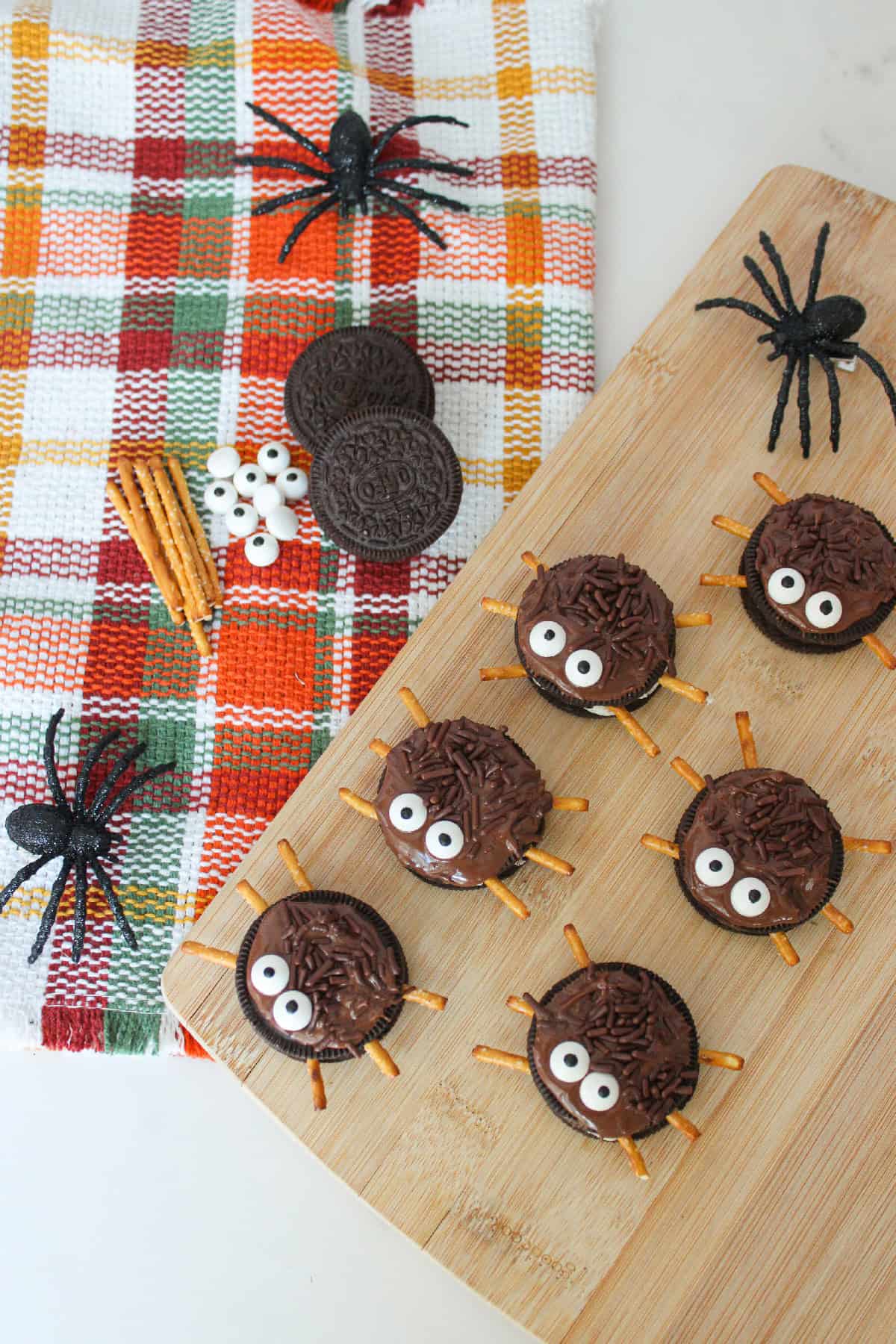 Fun Halloween Oreo spider treats with candy eyes and pretzel legs on a wooden board and plaid cloth.