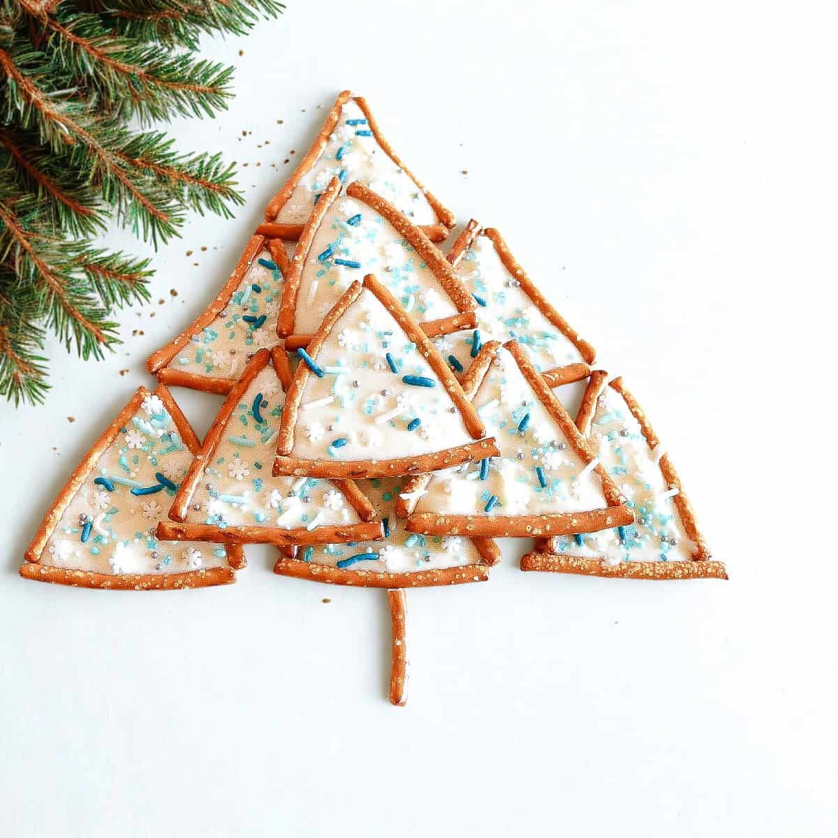 Pretzel triangles decorated with white chocolate and sprinkles arranged in the shape of a Christmas tree on white background with pine branch.