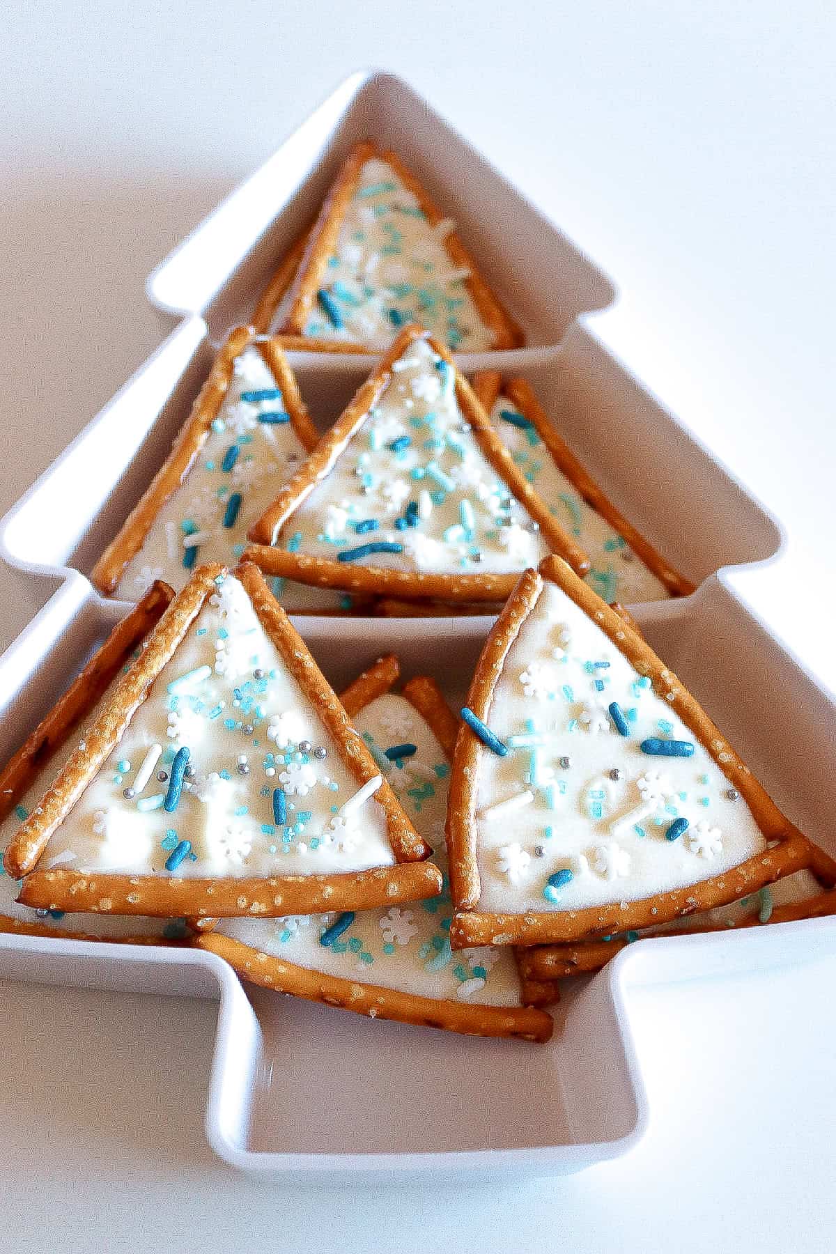 White Christmas tree-shaped dish filled with decorated pretzel triangle cookies topped with white chocolate and blue sprinkles.