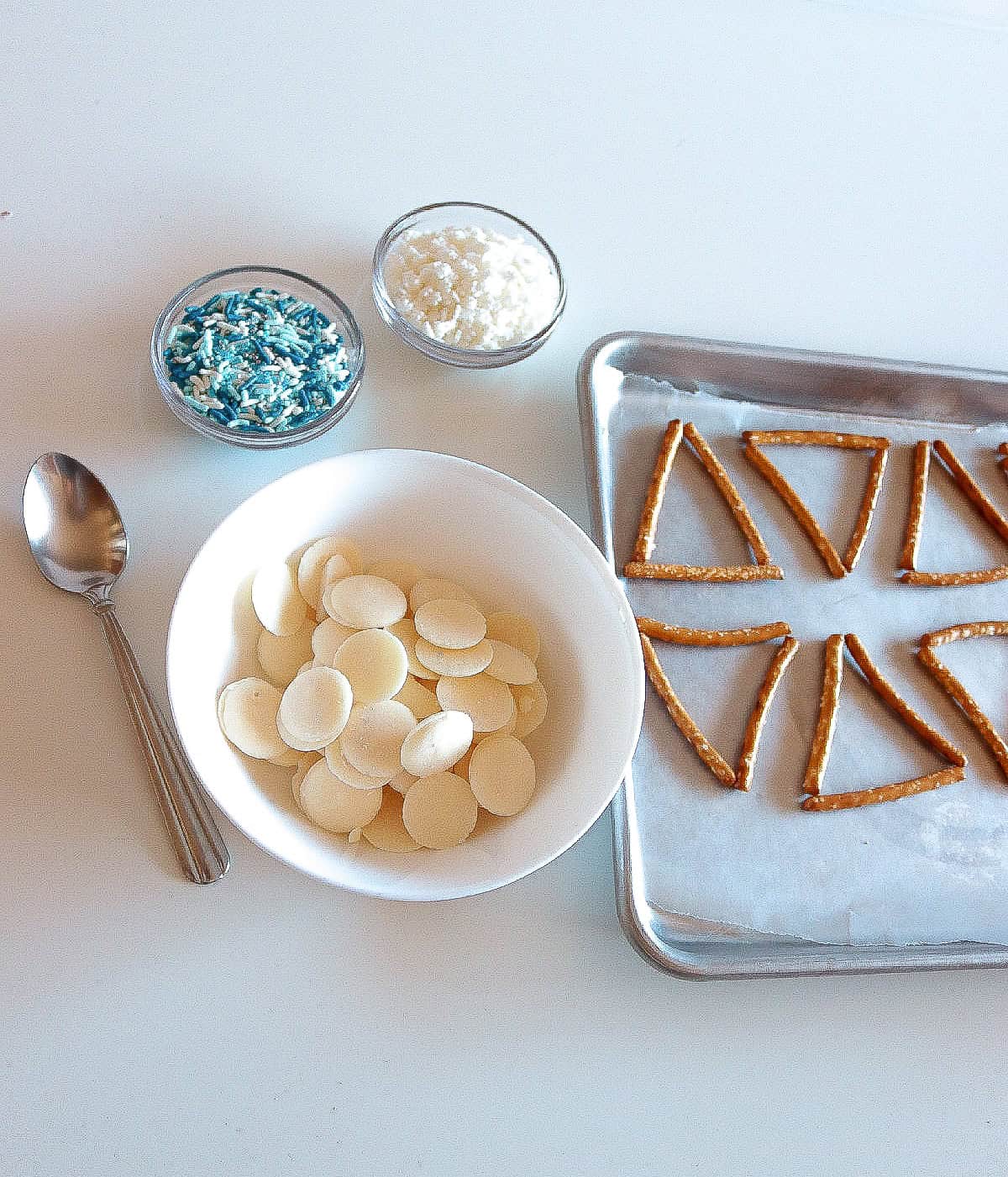 Baking tray with triangle-shaped pretzel frames, a bowl of white chocolate melts, spoon, and small bowls of blue sprinkles and crushed candy.