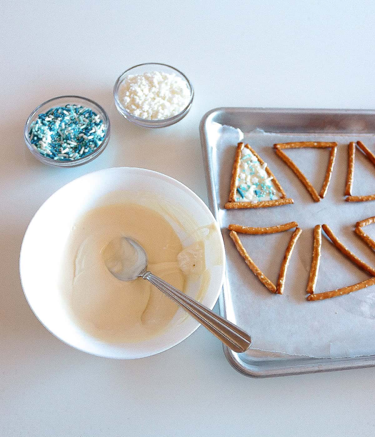 Melted white chocolate in a bowl with spoon, pretzel triangles on a baking tray, one decorated with white chocolate and blue sprinkles.