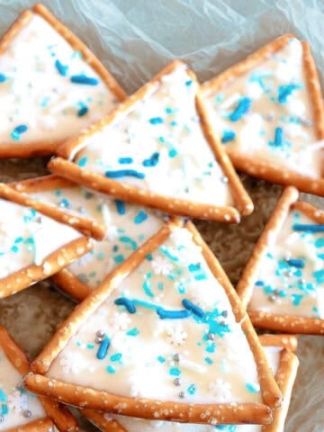 Triangle pretzel Christmas tree cookies decorated with white icing, blue sprinkles, and snowflake candy on parchment paper.