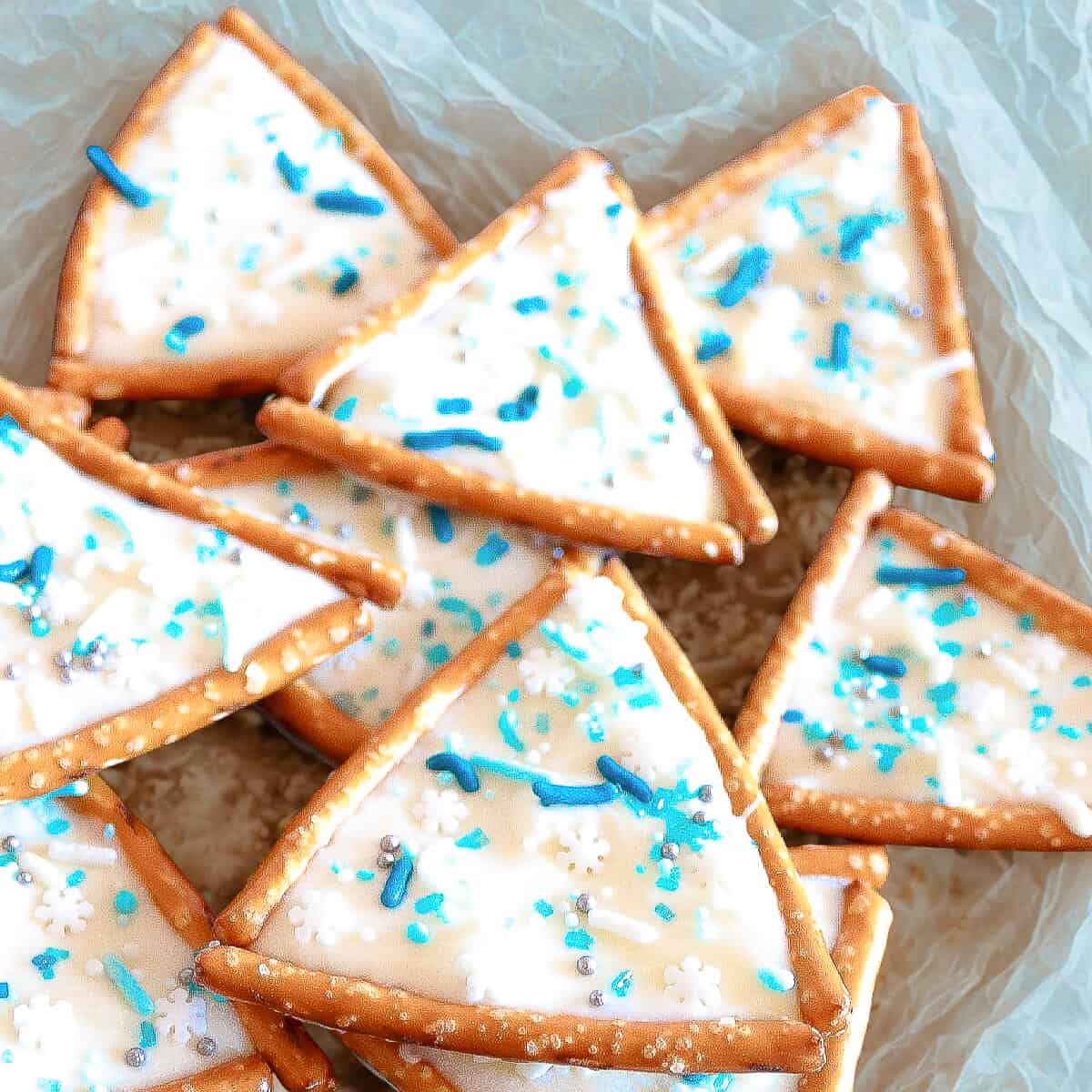 Triangle pretzel Christmas tree cookies decorated with white icing, blue sprinkles, and snowflake candy on parchment paper.