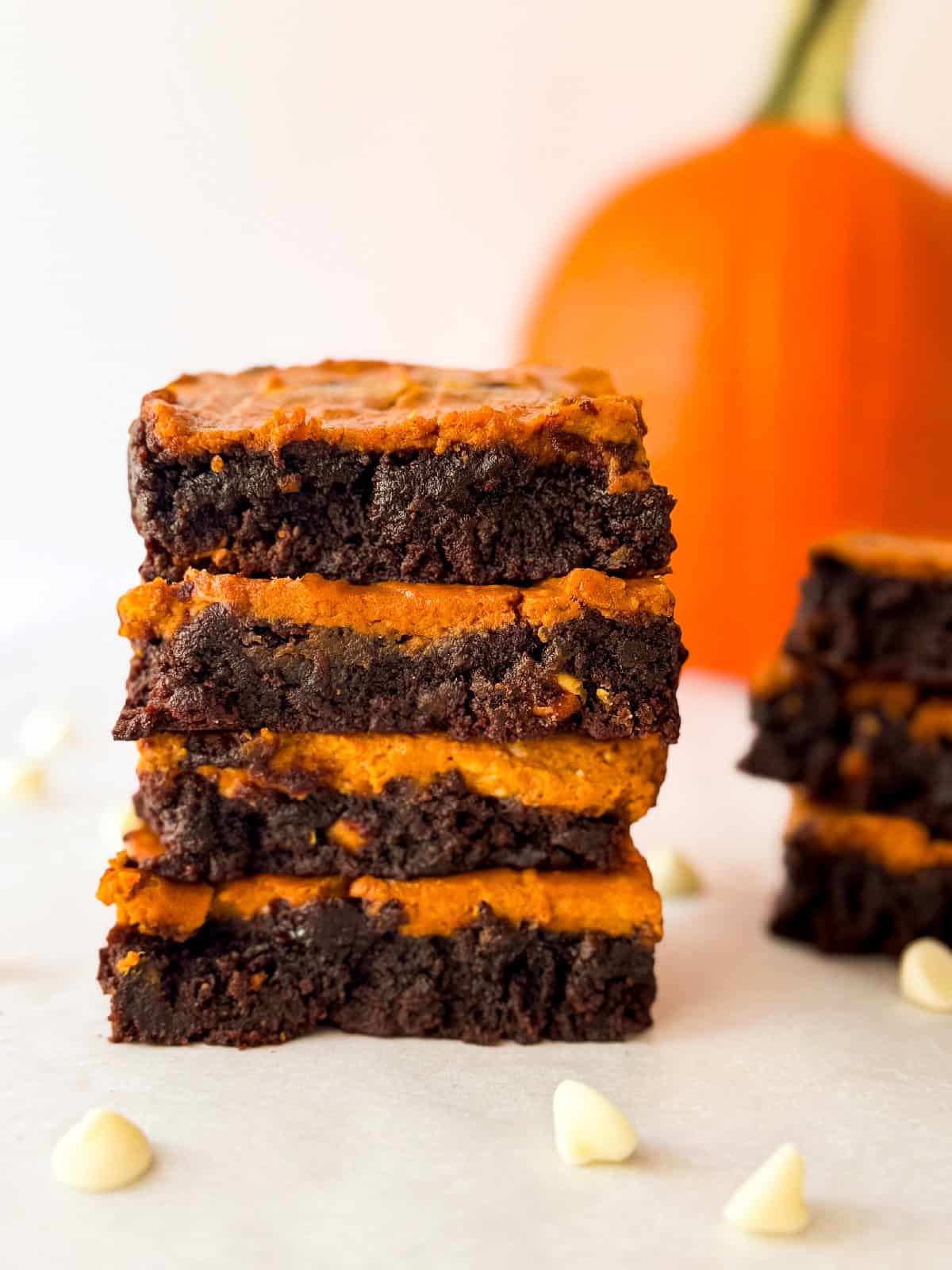 Stack of homemade pumpkin cheesecake brownies with rich chocolate layers and creamy pumpkin filling, styled with white chocolate chips on parchment and a blurred pumpkin in the background.