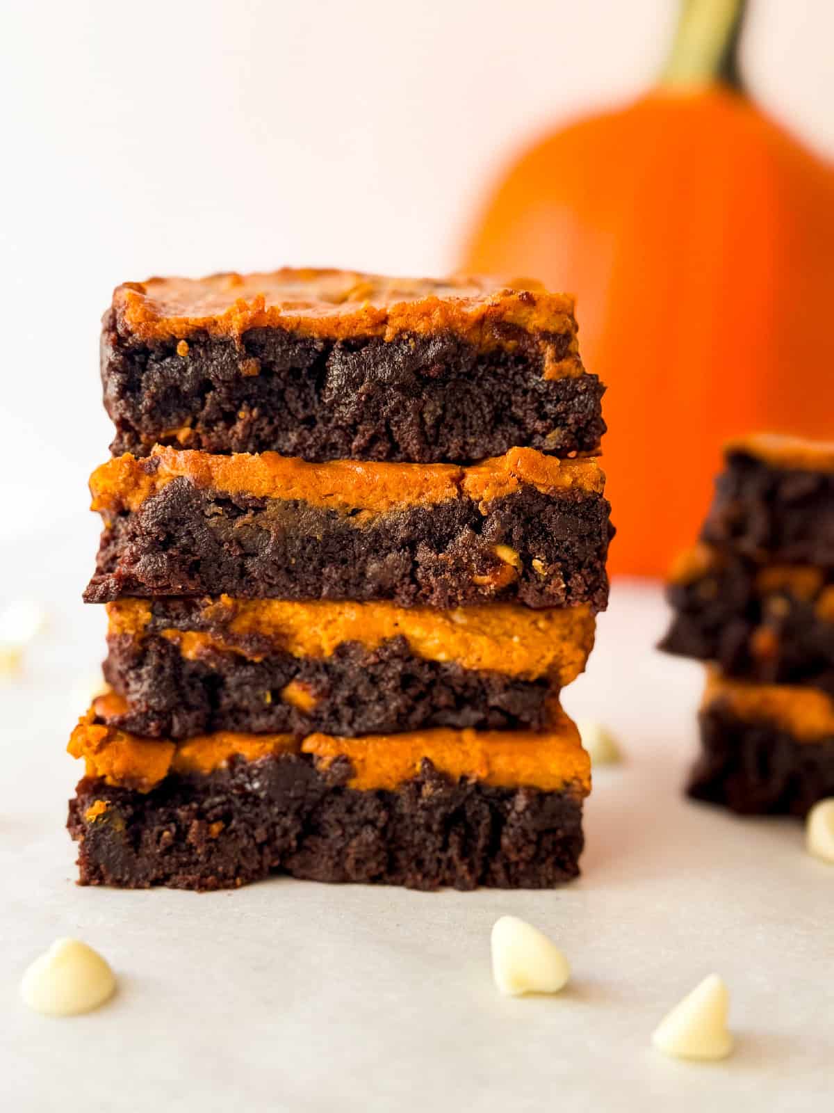 Close-up of stacked pumpkin cheesecake brownies showing fudgy chocolate base and bright pumpkin layer with scattered white chocolate chips and pumpkin in the background.