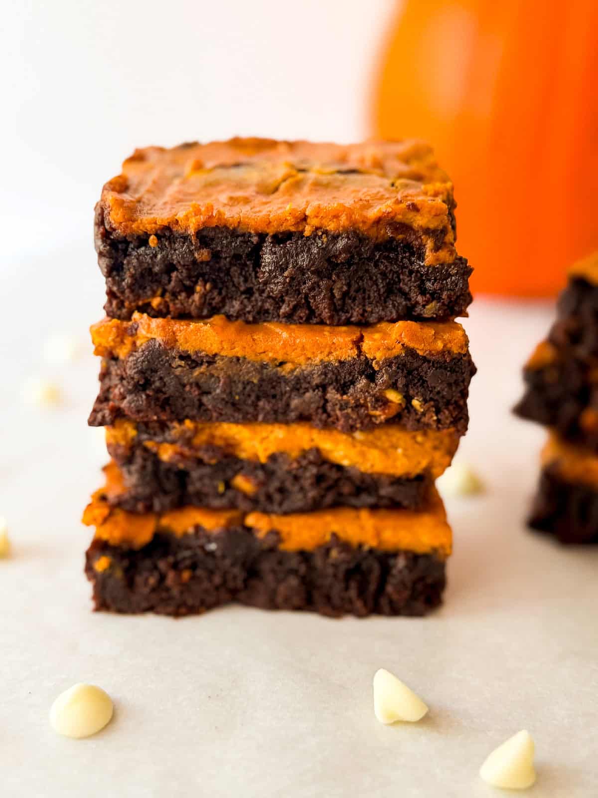 Tall stack of pumpkin cheesecake brownies highlighting the swirled chocolate and pumpkin layers on parchment paper with white chocolate chips and a blurred pumpkin behind.